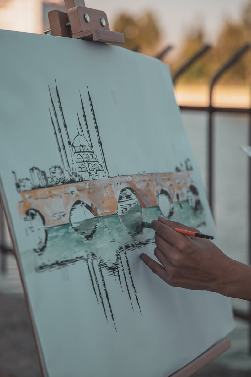 A hand painting a scenic bridge and dome on an easel, outdoors.