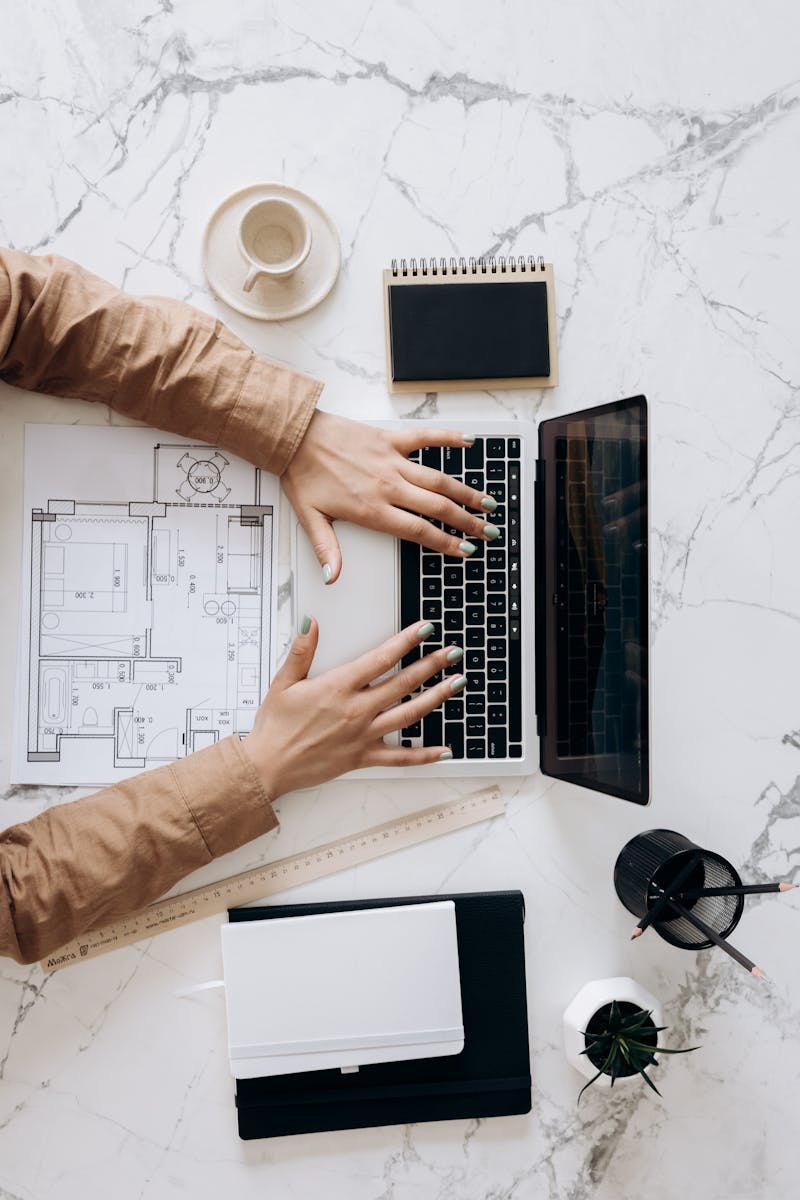 Overhead view of architect using laptop with blueprints and coffee on marble desk.