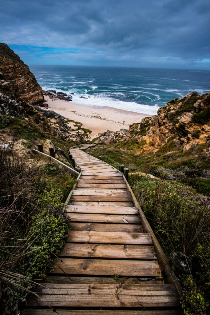Wooden stairs leading down to a scenic beach surrounded by cliffs under a dramatic sky.
