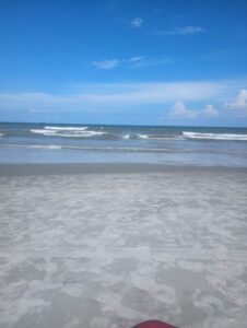 A wide, vertical shot of a quiet sandy beach looking out at the ocean under a bright blue sky. Gentle white waves roll onto the shore, and the foreground shows footprints scattered across the wide, gray sand.