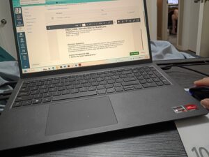 A close-up, high-angle shot of a black Windows laptop open on a desk, displaying an 'Introduction to Healthcare' assignment on the Eastern Florida State College Canvas interface. A hand is visible on the right, operating a computer mouse, while the screen shows coursework centered on analyzing medical errors and root cause analysis in a clinical setting.